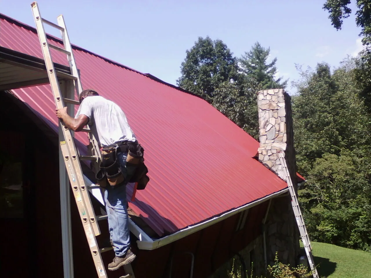 Roofer on a ladder beside a red metal roof and stone chimney — Soffit & Fascia Repair by Roofing Brothers in Irvine
